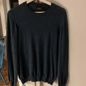 Mörkblå stickad tröja från Oscar Jacobson - En stilren mörkblå stickad tröja från Oscar Jacobson. Tröjan har rund halsringning, långa ärmar och ribbade muddar vid ärmslut och nederkant. Perfekt för lager-på-lager och enkel att matcha med jeans eller chinos. Tröjan är i nyskick, knappt använd. Bara att skicka om du har funderingar.
