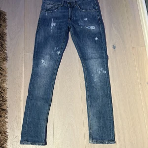 Dondup George jeans - Snygga blå Dondup George jeans i storlek 29. Jeansen har slitna detaljer och färgstänk för en cool, trendig look. Klassisk femficksmodell med dragkedja och knapp. Perfekta för dig som gillar en smal passform och Grisch stil. Dom har blivit lagade vi en av slitningarna, (se bild 5).