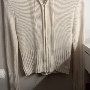 Vit ribbstickad hoodie med dragkedja - Ribbstickad vit tröja med dragkedja framtill och huva. Tröjan har långa ärmar och ribbad kant nedtill och vid ärmsluten. Perfekt på vintern och chill dagar. Använd 1 gång!