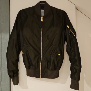 Svart bomberjacka Alpha Industries M - Snygg svart bomberjacka från Alpha Industries med guldfärgade dragkedjor och knappar. Jackan har klassisk ribbad krage, muddar och midja samt fickor med tryckknappar och en ärmficka med dragkedja. Tillverkad i slitstark polyester, perfekt för dig som gillar streetstyle.