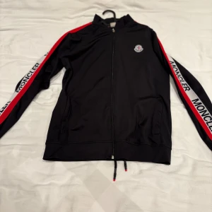 Svart Moncler trackoverall med röd/vit detalj - Svart trackjacka från Moncler med dragkedja framtill och ståkrage. Ärmarna har breda rödvita band med Moncler-logga längs hela längden. Jackan har två fickor och justerbar dragsko nertill. Perfekt för en sportig och trendig look. Byxor ingår!!!