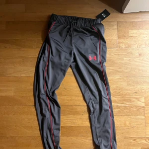 Under Armour svarta träningsbyxor - Snygga svarta träningsbyxor från Under Armour med röda detaljer längs benen och logga på låret. Byxorna har elastisk midja med snörning och är tillverkade i ett mjukt, syntetiskt material som andas. Perfekta för gymmet eller löprundan.