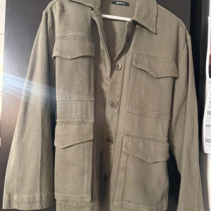 Grön jacka från Gina Tricot - Stilren grön jacka från Gina Tricot med fyra stora fickor framtill, knäppning med knappar och bälte i midjan. Jackan har klassisk krage och raka ärmar, perfekt för lager-på-lager och en chill look. Jackan har även slits på sidan.