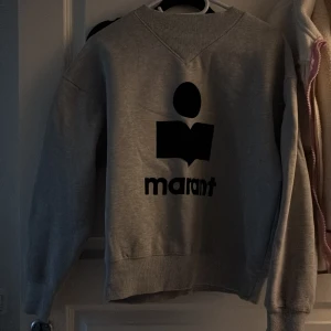 Isabel Marant tröja  - Säljer en grå sweatshirt från Isabel Marant Étoile med svart logga och tryck på bröstet. Tröjan har rund hals, ribbade muddar och är tillverkad i mjuk bomullsmix. Perfekt för en chill och stilren look.