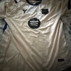 Barcelona Lamine Yamal bortatröja - Säljer en beige Barcelona bortatröja med Lamine Yamal och nummer 10 på ryggen. Tröjan har svarta detaljer, blå inslag på sidorna och klassiska klubbmärket framtill. Tillverkad i lätt och ventilerande material, perfekt för fotboll eller att bära till vardags.