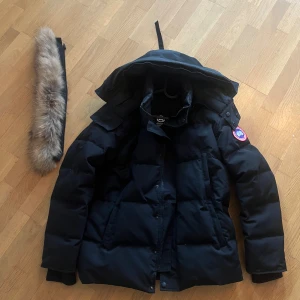 Svart dunjacka från Canada Goose Wyndham - Säljer en svart dunjacka från Canada Goose med avtagbar huva och pälskant. Jackan har dragkedja och knappar framtill samt två stora fickor. Perfekt för kalla vinterdagar och riktigt varm. Säljer då den inte används längre.