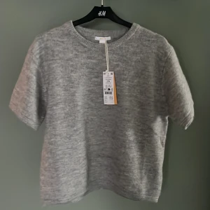 Grå stickad tröja från Gina Tricot - Mysig grå stickad tröja från Gina Tricot med kort ärm och rund halsringning. Tröjan har en enkel och stilren design, perfekt för lager-på-lager och passar till många olika outfits. Materialet är mjukt och ger en avslappnad känsla.