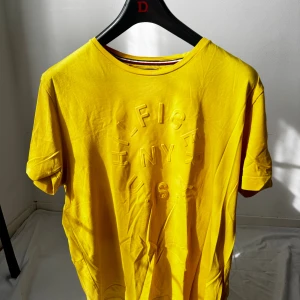 Gul t-shirt Tommy Hilfiger XL - Gul t-shirt från Tommy Hilfiger i ekologisk bomull. T-shirten har korta ärmar och ett upphöjt tryck med texten 'HILFIGER NY U.S.A.' på bröstet. Klassisk rund halsringning och snygga detaljer vid kragen.