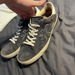Premiata skor - Snygga svarta sneakers från Premiata i mocka med vita detaljer och snörning. Skorna har en klassisk låg profil, vit sula och rund tå. Insidan är fodrad i ljust skinn och yttersulan är svart med Premiata-logga. 
