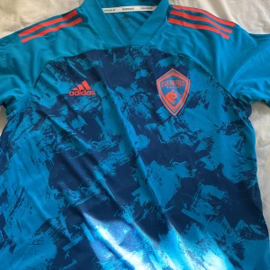 Colorado Rapids blå fotbollströja Adidas - Snygg Colorado Rapids fotbollströja från Adidas i blått med mörkblått abstrakt mönster och orange detaljer. Kortärmad modell med tre orange ränder på axlarna och klubbmärke på bröstet. Tillverkad i lätt och ventilerande Primeblue-material.