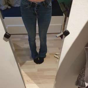 Blå bootcut jeans med hög midja - Säljer ett par klassiska blå jeans med bootcut-snitt och hög midja. Jeansen har fem fickor, bälteshällor och stängs med dragkedja och knapp. Materialet är jeans i bomullsmix och färgen är en snygg mellanblå ton. Perfekta för dig som gillar en tidlös look.