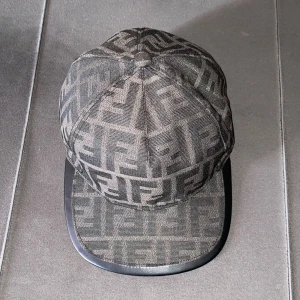 Fendi keps med FF-mönster - Snygg keps från Fendi med det klassiska FF-logomönstret i mörkgrå och svart ton. Kepsen har böjd skärm med svart kant och justerbar rem bak. Tillverkad i bomull och polyester för en bekväm passform. Perfekt för dig som vill ha en lyxig streetwear-look.