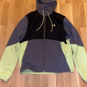 Under Armour vindjacka med huva - Snygg vindjacka från Under Armour i svart, grått och ljusgrönt. Jackan har huva med snörning, hel dragkedja och logga på bröstet. Perfekt för dig som gillar sportig stil och vill ha något lätt och funktionellt.