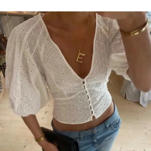 Vit broderad blus med puffärm - Superfin vit blus med broderade hål och puffiga trekvartsärmar. Djup v-ringning och knappar framtill ger en romantisk vibe. Croppad passform som passar perfekt till jeans eller kjol. Perfekt för vår och sommar!