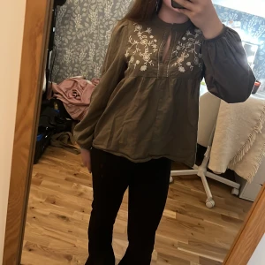 Olivgrön blus med broderade blommor - Olivgrön blus med vita broderade blommor över bröstet och en liten v-ringning framtill. Blusen har långa puffiga ärmar med resår vid handlederna och är tillverkad i ett mjukt bomullstyg. Den har en lös och flowy passform som ger en chill vibe.