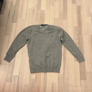 Grå v-ringad tröja från Ralph Lauren - Snygg grå v-ringad tröja från Ralph Lauren med diskret broderad logga på bröstet. Tröjan har lång ärm och ribbade muddar vid ärmslut och nederkant. Perfekt för lager-på-lager och enkel att matcha med jeans eller chinos.