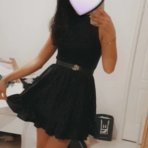 Svart glittrig kortklänning - Supersnygg svart glittrig kortklänning med markerad midja. Klänningen har ärmlös design och lätt utsvängd kjol som ger en feminin siluett. Perfekt för dig som vill sticka ut med stil och glitter. 