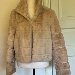 Beige pälsjacka från Guess med pärldetaljer - Supermjuk och fluffig beige pälsjacka från Guess med bred krage och horisontella sömmar. Jackan hart långa ärmar med snygga pärldetaljer och band längs armarna. Perfekt för dig som vill sticka ut med en lyxig och trendig look.