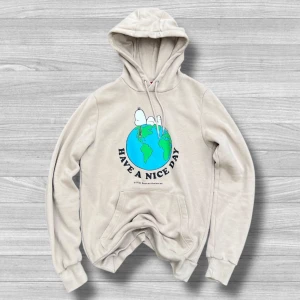 Beige Peanuts hoodie med Snoopy XS - Beige hoodie från Peanuts med tryck av Snoopy som chillar på en jordglob och texten 'Have a nice day'. Tröjan har huva med snörning och känguruficka framtill. Perfekt för dig som gillar avslappnad stil och ikoniska motiv.