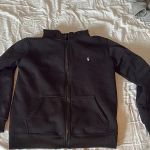 Svart zip hoodie från Polo Ralph Lauren - Svart hoodie från Polo Ralph Lauren med dragkedja framtill och två stora fickor. Klassisk design med broderad logga på bröstet. Mjuk insida och ribbade muddar vid ärmslut och nederkant. Perfekt för chill dagar eller när du vill ha en clean look. Storlek medium men kan passa s också 
