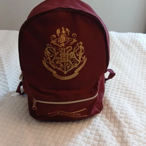 Vinröd Hogwarts ryggsäck med gulddetaljer - Säljer en vinröd ryggsäck med Hogwarts-emblem i guld på framsidan. Väskan har ett stort huvudfack, en mindre ficka framtill med dragkedja och justerbara axelremmar. Perfekt för dig som älskar Harry Potter och vill ha en snygg och praktisk väska till skolan eller stan.
