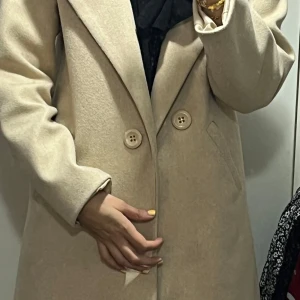 Beige kappa med knappar - Snygg beige kappa med två stora knappar framtill och klassisk krage. Jackan har raka fickor på sidorna och är helfodrad. Perfekt för dig som vill ha en stilren och tidlös look till höst och vinter.