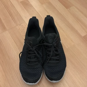 Svarta Nike sneakers med vit sula - Säljer ett par svarta Nike sneakers med vit sula och klassisk Nike-logga på sidan. Ovandelen är i mesh-liknande syntetmaterial för bra andningsförmåga. Skorna har snörning och en sportig, modern look som passar perfekt till streetwear.