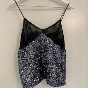 Glittrig svart och silver paljettopp - Snygg glittrig topp med tunna axelband och transparent mesh upptill. Nedre delen är täckt av silverfärgade paljetter som ger ett riktigt party-vibe. Toppen är ärmlös och har en loose passform, perfekt för att sticka ut i mängden.