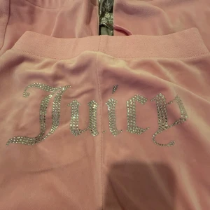 Juicy Couture rosa velourbyxor - Rosa mjukisbyxor från Juicy Couture i velour med bred resår i midjan och snörning framtill. Baktill står det 'Juicy' i glittrande strasstenar. Byxorna har raka ben och en liten logga framtill. Perfekta för en chill och trendig look. Inga stenar har lossnat och de är inte trasiga längst ner🩷