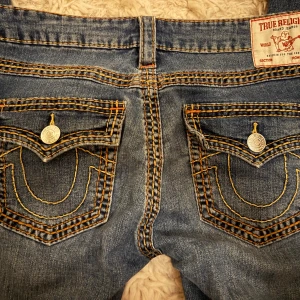 True Religion Joey jeans med bootcut - Säljer ett par blå True Religion Joey jeans med bootcut och låg midja. Jeansen har tydliga kontrastsömmar i orange och silverfärgade knappar på fickorna. Klassiska bakfickor med True Religion-logga och grov denimkänsla. Perfekta för dig som gillar statement-jeans.