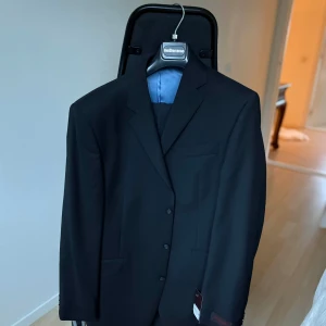 Svart kostym – Pierre Cardin Paris (stl 50) - Stilren svart kostym från det klassiska modemärket Pierre Cardin. En tidlös design med snygg passform och elegant ljusblått innerfoder. Perfekt för både formella tillfällen och fest.  Storlek: 50  Färg: Svart  Märke: Pierre Cardin Paris  Material: 100% ull  Nypris: ca 3995 kr  Skick: Helt ny med tags kvar
