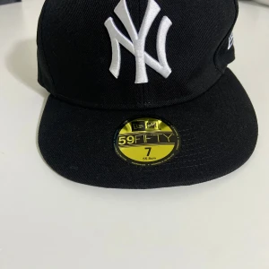 Svart New Era Yankees keps 59FIFTY - Snygg svart New Era 59FIFTY keps med klassiskt New York Yankees broderat vitt logotyp framtill. Platt skärm med ljus undersida och MLB-logga baktill. Perfekt för dig som gillar streetstyle och baseball. Kepsen är i fast modell och har coola detaljer invändigt.