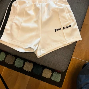 Vita shorts från Palm Angels med svarta ränder - Snygga vita shorts från Palm Angels med svarta ränder på sidorna och logga broderad framtill. Elastisk midja och sportig design, perfekta för sommaren.