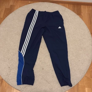 Adidas blå träningsbyxor med ränder - Mörkblå träningsbyxor från Adidas med klassiska vita ränder längs ena benet och broderad logga på låret. Byxorna har elastisk midja och en blå detalj nertill på benet. Perfekta för sport eller chill.
