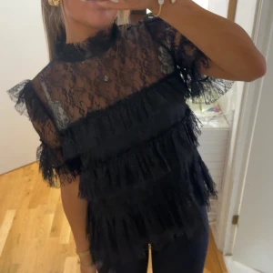 Svart spetsblus med volanger - Supersnygg svart blus i spets med volangdetaljer och korta ärmar. Transparent överdel med hög hals och flera lager volanger som ger en cool och trendig look. Perfekt att styla med jeans eller kjol för en extra touch. 1800kr ny