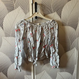  blommig offshoulder blus - Säljer en söt offshoulder blus med tunna blå och vita ränder och små röda blommor. Blusen har långa ärmar och knytning nertill för en avslappnad vibe. Perfekt för dig som gillar romantisk och trendig stil.