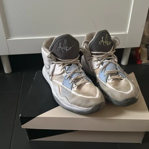 Nike Kyrie basket sneakers grå/vit - Säljer ett par Nike Kyrie basket sneakers i grått och vitt med ljusblå detaljer. Skorna har högt skaft, snörning och coola signaturdetaljer på plösen och hälen. Snygg design med svart Swoosh och meshpartier för extra ventilation.