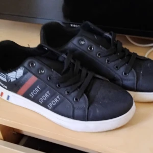 Svarta sneakers Sport - Svarta sneakers med vita sulor och svarta snören. Sidan har röda och grå ränder samt texten 'SPORT' tryckt tre gånger. Dekorativt vitt mönster vid hälen och små franska flaggdetaljer på sulan. Perfekta för en sportig och avslappnad stil.