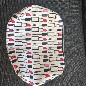 Vit necessär med läppstiftsmönster - Säljer en söt vit necessär med färgglatt läppstiftsmönster i rött, rosa och lila. Väskan har dragkedja upptill och är tillverkad i syntetmaterial med lätt glansig yta. Perfekt för smink eller småprylar i väskan.