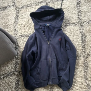Marinblå hoodie från Polo Ralph Lauren -  Xl barn Snygg marinblå hoodie från Polo Ralph Lauren med dragkedja och huva. Klassisk röd broderad logga på bröstet och stora fickor framtill. Perfekt för chill dagar och enkel att matcha med jeans eller joggers.