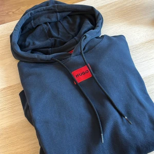 Svart hoodie från Hugo med röd logga - Svart hoodie från Hugo med en snygg röd logotyp på bröstet. Tröjan har huva med snörning och är tillverkad i mjuk bomull. Perfekt för en chill och stilren look.