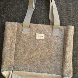 Beige shoppingväska från Marella - Stilren shoppingväska från Marella i beige filtmaterial med breda, matchande handtag. Väskan har en fyrkantig form, detaljer i syntet längst ner och ett rymligt innerfack med dragkedja. Perfekt för dig som vill ha en enkel men snygg väska till vardagen.