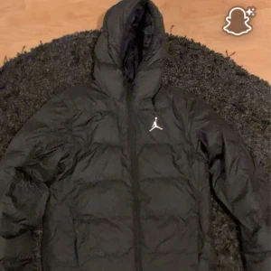 Svart pufferjacka från Jordan - Svart pufferjacka från Jordan med huva och klassisk Jumpman-logga på bröstet. Jackan har dragkedja framtill och är quiltad för extra värme. Perfekt för kalla dagar och har en clean, sportig look som funkar till streetwear.