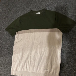 Tvåfärgad t-shirt från SAMS - Snygg t-shirt från SAMS i storlek S med olivgrön överdel och beige nedre del. Klassisk rund halsringning och korta ärmar. Perfekt för dig som gillar stilrena färgblock och enkel design.