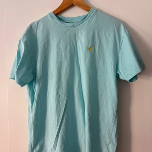 Ljusblå t-shirt från Polo Ralph Lauren - Snygg ljusblå t-shirt från Polo Ralph Lauren med klassisk gul broderad logga på bröstet. T-shirten har rund halsringning och korta ärmar, perfekt för en chill och stilren look. Tillverkad i mjuk bomull som känns skön mot huden.