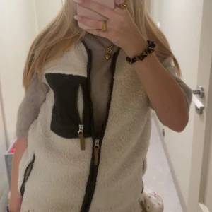 Vit fleeceväst med svarta detaljer - Mysig och trendig vit fleeceväst med svarta detaljer och dragkedjor. Västen har en cool bröstficka med dragkedja och kontrasttyg, samt två sidofickor. Perfekt att slänga över en hoodie eller tröja för extra värme och stil.