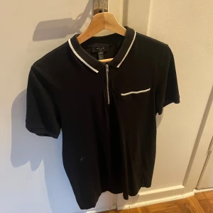 Svart t-shirt med dragkedja från New Look - Svart t-shirt från New Look med vit kant på kragen och fickan. Dragkedja framtill och korta ärmar ger en clean och modern vibe. Perfekt för dig som gillar stilrena detaljer och vill ha något som sticker ut lite extra.