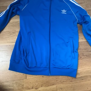 Blå adidas tröja  - Säljer en klassisk blå Adidas vindjacka med vita ränder längs ärmarna och vit logga på bröstet. Jackan har dragkedja framtill, två fickor och ribbade muddar. Perfekt för dig som gillar sportig och retro stil. Ny så kostar den 1000kr
