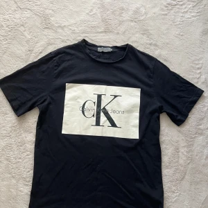 Svart t-shirt Calvin Klein Jeans - Svart t-shirt från Calvin Klein Jeans med stor vit logga på bröstet. Klassisk rund hals och korta ärmar. T-shirten är gjord i mjuk bomull och har en normal passform. Perfekt för dig som gillar stilrena och enkla plagg med tydlig branding.