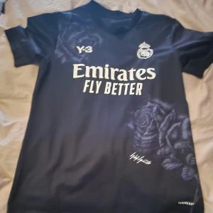 Real Madrid x Y-3 fotbolls tshirt - Snygg svart Real Madrid fotbollströja från Y-3 med vita tryck, klubbmärke och Emirates Fly Better-logga. Unik rosdetalj i mönstret både fram och bak. Tillverkad i lätt och ventilerande material, perfekt för match eller träning.